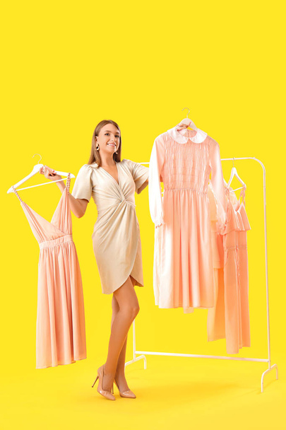 Beautiful happy young woman and rack with different stylish clothes on yellow background - Photo, Image
