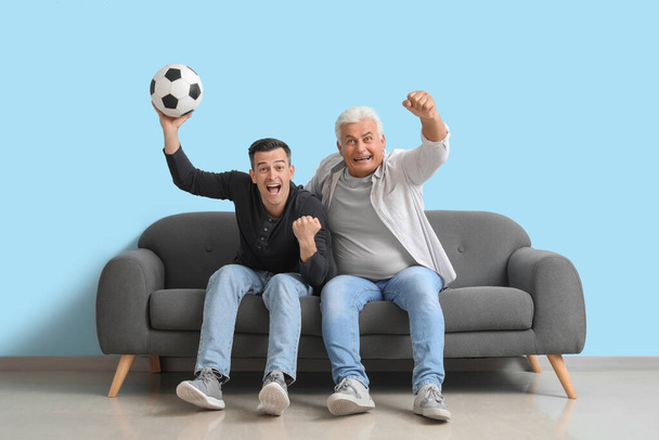 Jovem com seu pai assistindo jogo de futebol no sofá perto da parede azul - Foto, Imagem