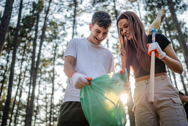 Çift ya da genç arkadaşlar Z erkek ve beyaz kadın atık plastik şişeleri ve kağıtları toplayarak güneşli bir günde çevre koruma kavramının doğasını temizlerler. - Fotoğraf, Görsel