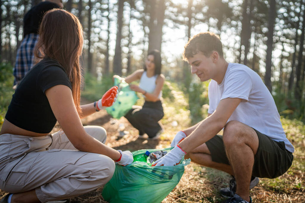 group of teenage friends gen Z male and female caucasian men women picking up waste garbage plastic bottles and paper from the forest cleaning up nature in sunny day environmental care ecology concept - Photo, Image