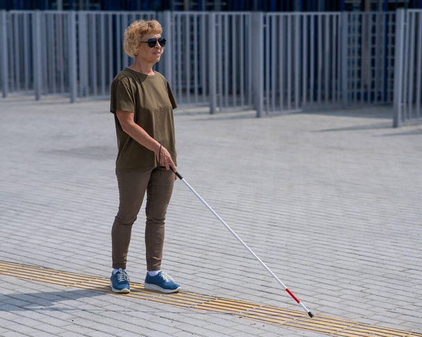 An elderly blind woman walks with a cane along a tactile tile - Фото, изображение