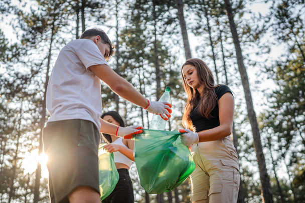 group of teenage friends gen Z male and female caucasian men women picking up waste garbage plastic bottles and paper from the forest cleaning up nature in sunny day environmental care ecology concept - Photo, Image