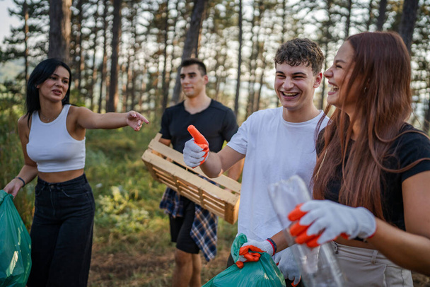 grupa nastolatków przyjaciele gen Z mężczyzna i kobieta kaukaski mężczyźni kobiety zbieranie śmieci plastikowe butelki i papier z lasu sprzątanie przyrody w słoneczny dzień ekologia opieki koncepcja - Zdjęcie, obraz
