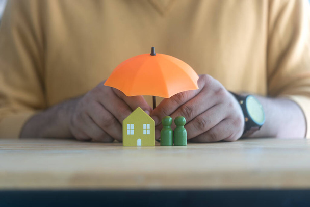 Чоловік тримає помаранчеву парасолькову кришку дерев'яної домашньої моделі та дерев'яної людської моделі. Захист сім'ї. Концепції страхування - Фото, зображення