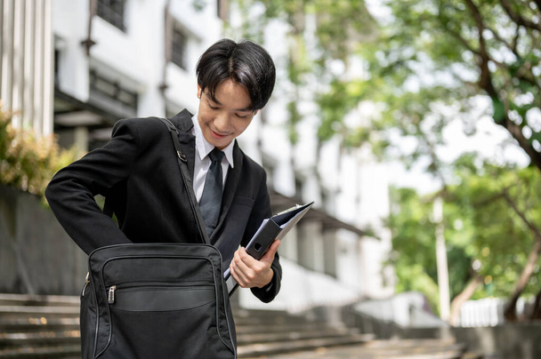 Un joven empresario asiático feliz y guapo o un empleado de oficina masculino en un traje formal de negocios encuentra su teléfono en su maletín mientras baja las escaleras de la oficina. - Foto, imagen