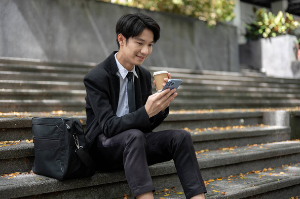 Um jovem empresário asiático feliz e bonito ou funcionário de escritório masculino está usando seu smartphone para verificar suas mensagens e tomar café enquanto está sentado em uma escadaria em frente ao prédio. - Foto, Imagem