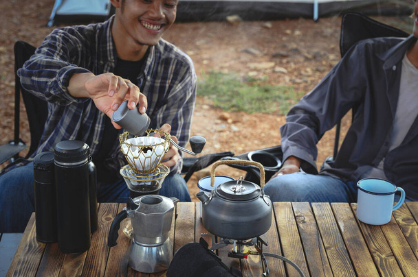 Asian young man two persons sitting enjoy making drip coffee drink near a camping tent, coffee drink relaxing on the morning summer camping vacation. - Photo, Image