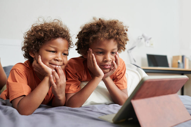 Usmívající se děti sledující kreslené filmy na tabletu počítače při relaxaci na posteli - Fotografie, Obrázek