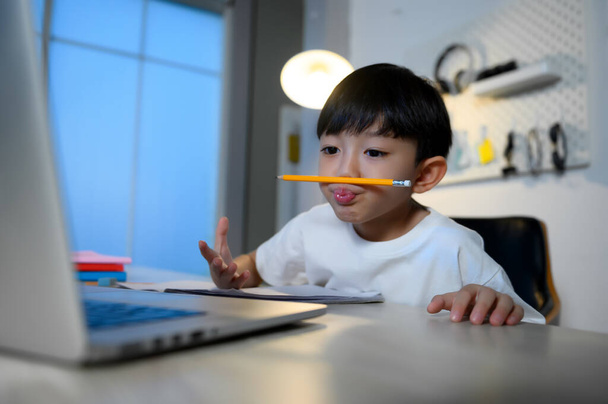 Azjatycki chłopiec trzyma ołówek między ustami i nosem podczas studiów i koncentruje się na pracy domowej i uczenia się sam w pokoju w domu z narzędzi cyfrowych Laptop. Edukacja Asian Kids Concept. - Zdjęcie, obraz