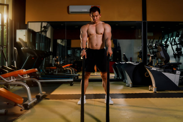 Gespierde bodybuilder sterke jonge Aziatische man training met strijd touw oefening in de fitnessruimte. Fitness training spieropbouw of bodybuilding met battle rope apparatuur. - Foto, afbeelding