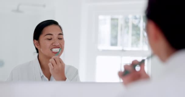 Čištění zubů, zrcadlo a žena v domácí koupelně, zdraví a wellness v domě. Zubní, zubní kartáček čištění a odraz asijské osoby pro ústní péči, ústní hygienu a zubní pastu v dopoledních hodinách. - Záběry, video