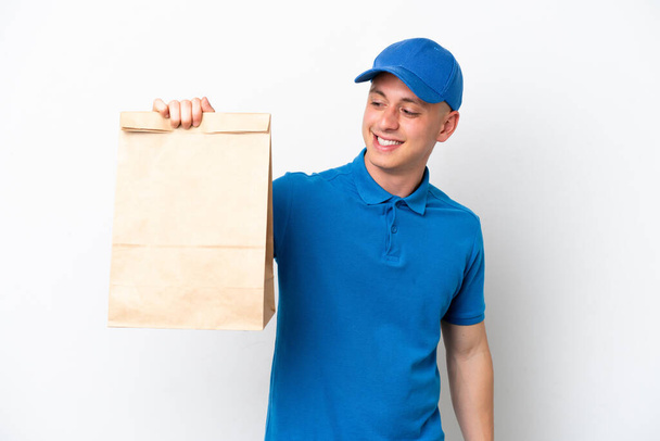 Joven brasileño llevando una bolsa de comida para llevar aislada sobre fondo blanco mirando hacia un lado y sonriendo - Foto, Imagen