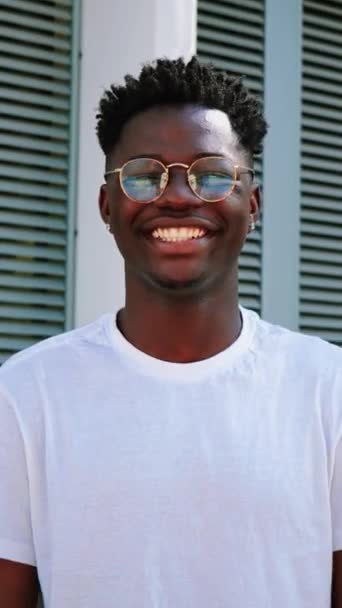 Κάθετο πορτραίτο ενός νεαρού Αφροαμερικανού εφήβου που χαμογελάει και κοιτάζει την κάμερα να στέκεται έξω. Μπροστά όψη του ευτυχισμένος και χαρούμενος άνθρωπος με γυαλιά ηλίου και λευκά τέλεια δόντια. αργή κίνηση. Υψηλή - Πλάνα, βίντεο