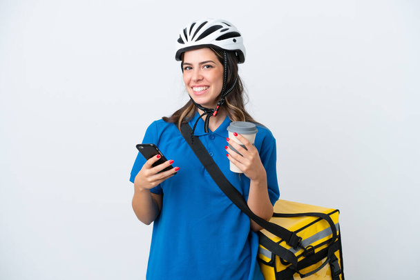 Young delivery woman with thermal backpack isolated on white background holding coffee to take away and a mobile - Fotó, kép