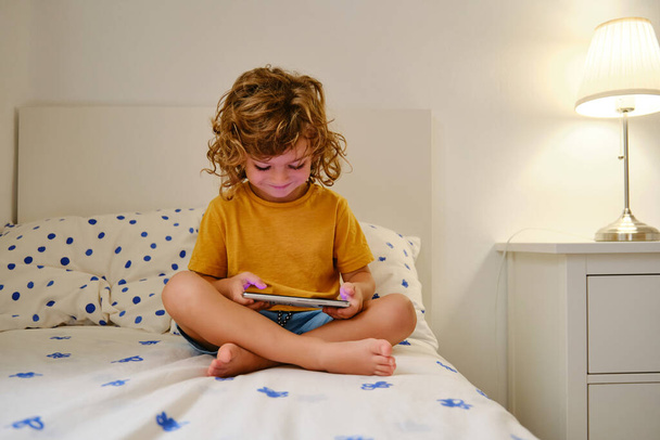 Celé tělo rozkošné kudrnaté vlasy chlapec v domácím oblečení sedí na posteli se zkříženýma nohama a hraje hru na mobilním telefonu na večerní čas - Fotografie, Obrázek