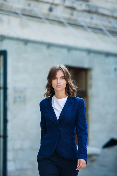 Muotokuva kauniista nuoresta naisesta, jolla on pitkät ruskeat hiukset, pukeutunut siniseen takkiin, seisoo toimistorakennuksen taustalla. vahva ja luottavainen liiketoiminnan nainen - Valokuva, kuva