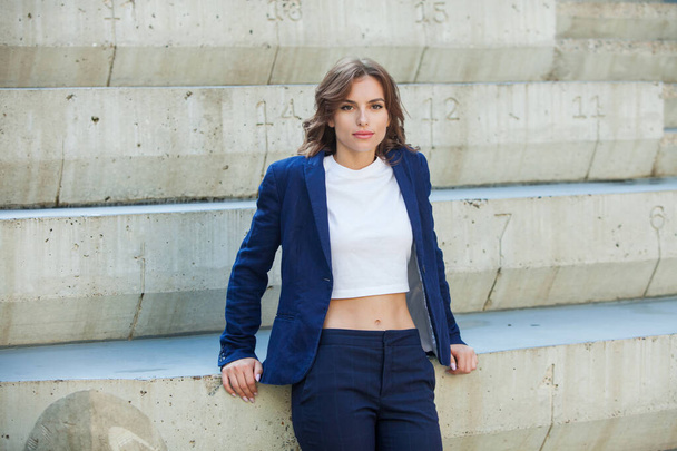 Portrait of a beautiful young woman with long brown hair, dressed in a blue jacket, standing against the background of an office building. strong and confident business woman - Photo, Image