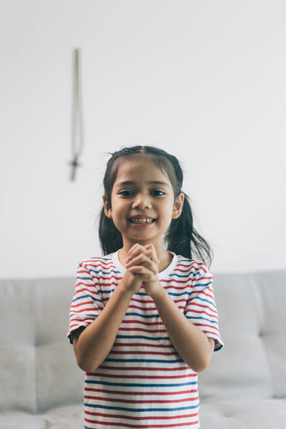 Egy kis ázsiai lány mosolyog és a kamerába néz, miközben Istenért imádkozik otthon a kanapén. - Fotó, kép