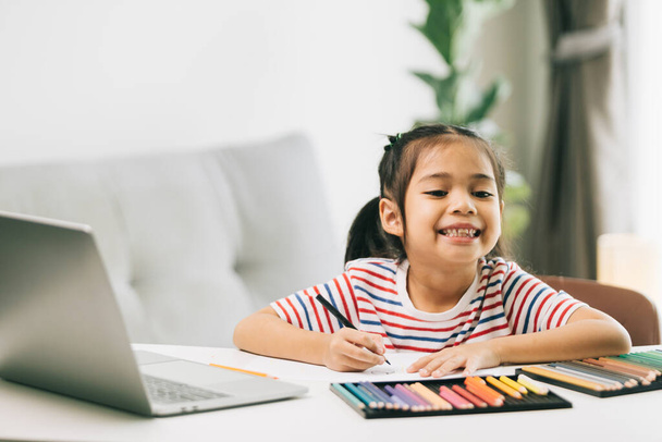 Evde renkli kalemler ve dizüstü bilgisayarlarla resim çizen Asyalı küçük kız. - Fotoğraf, Görsel