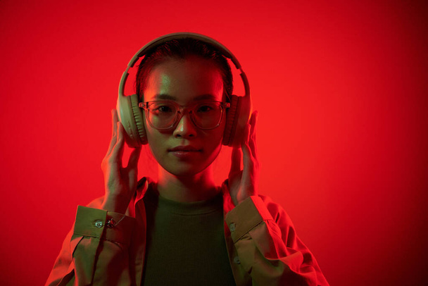 Portrét mladé asijské ženy nosí brýle a sluchátka a dívá se do kamery - Fotografie, Obrázek