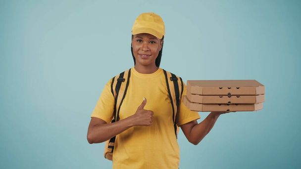 Portrait of delivery woman in yellow cap and tshirt holding stack of pizza boxes and shows a thumbs up gesture. Isolated on blue background. - 写真・画像