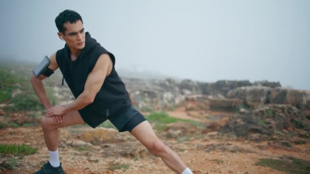 Active guy doing exercise lunges on cliff. Focused athletic male stretching legs prepare trail race competition at cloudy sky. Handsome strong runner warming up body listening music earphones on rocks - Footage, Video