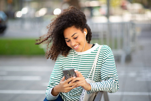 Портрет молодой улыбающейся женщины, сидящей с мобильным телефоном и слушающей наушники - Фото, изображение