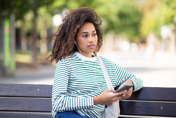 Portret jonge Afrikaanse Amerikaanse vrouw zittend op park bank met mobiele telefoon - Foto, afbeelding