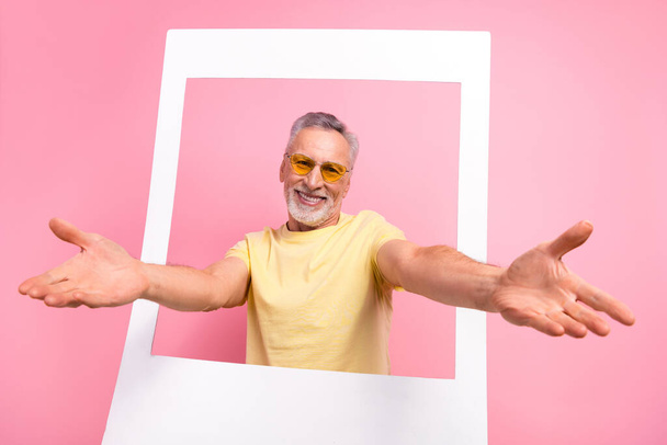 Photo of friendly cheerful man with beard dressed yellow t-shirt in sunglass stretching arms from frame isolated on pink color background. - Fotó, kép