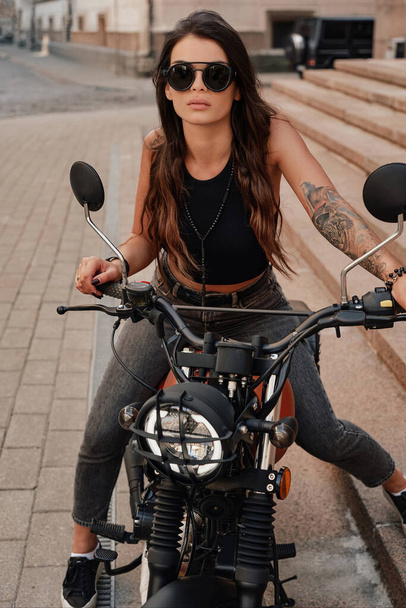 Κομψή μελαχρινή με τζιν και φανελάκι, με γυαλιά ηλίου και τατουάζ στο μπράτσο, ποζάρει πάνω στη ρετρό μοτοσικλέτα της σε ένα παλιό πλακόστρωτο δρόμο στην Ευρώπη - Φωτογραφία, εικόνα