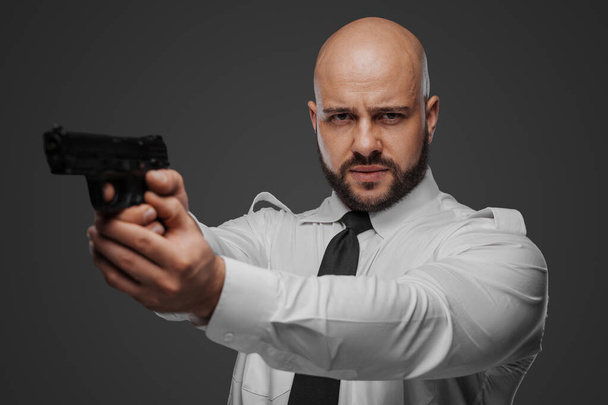 Φαλακρός, γενειοφόρος άνδρας με λευκή μπλούζα και γραβάτα στοχεύει ένα πιστόλι, διοχετεύοντας έναν ντετέκτιβ ή σωματοφύλακα vibe σε ένα γκρίζο σκηνικό στούντιο - Φωτογραφία, εικόνα