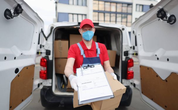 Courier driver in medical protective mask and gloves holds out documents with delivery receipt. Safe customer service for cargo delivery concept - Photo, Image