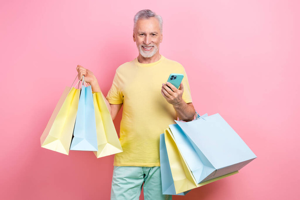 Photo of clever senior man with beard dressed yellow t-shirt hold new clothes make order in smartphone isolated on pink color background. - Foto, afbeelding