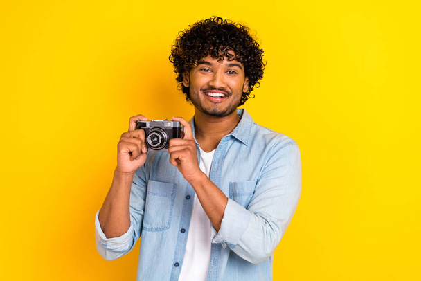 Photo of positive good mood nice man with wavy hairstyle wear jeans shirt hands hold retro camera isolated on yellow color background. - Photo, Image