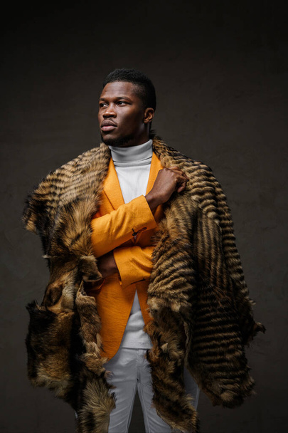 Portrait of a stylish dark-skinned man wearing a yellow jacket and a wild animal skin draped over his shoulders, posing against a dark background - Фото, изображение