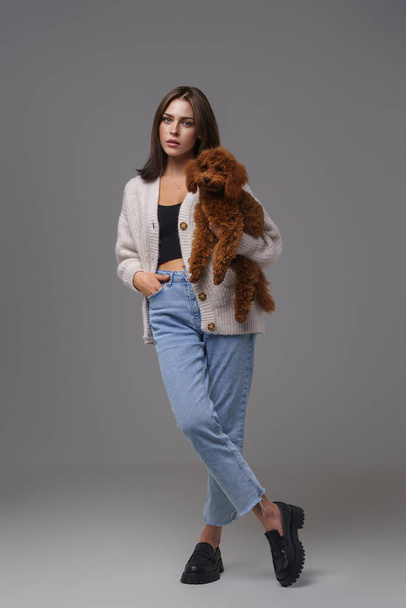 Ολόσωμο πορτραίτο μιας σαγηνευτικής μελαχρινής σε casual ένδυμα κρατώντας το καφέ σκυλάκι της σε γκρι φόντο στούντιο - Φωτογραφία, εικόνα