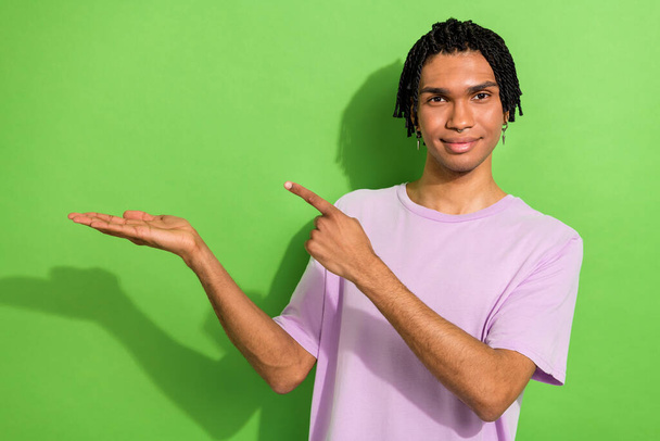 Photo of funny positive guy wear pink t-shirt showing finger holding arm empty space isolated green color background. - Foto, immagini