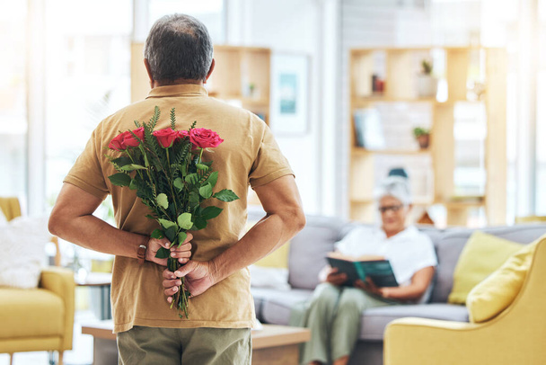 Liefde, verrassing en senior man met rozen achter de rug voor romantiek, dankbaarheid en toewijding aan vrouw in de woonkamer van huis. Paar, persoon en bloemen in de hand voor cadeau en huwelijksverjaardag. - Foto, afbeelding