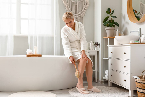 Mature woman massaging her leg with brush in bathroom - Фото, изображение
