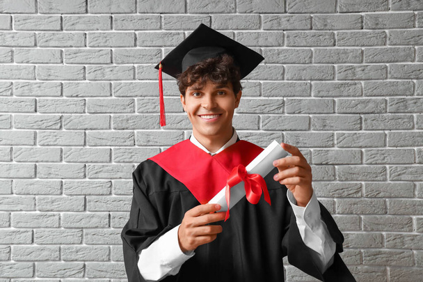 Male graduate student with diploma on grey brick background - Photo, Image