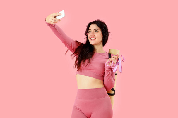 Sportowa młoda kobieta ze skakanką i matą do jogi robi selfie na różowym tle - Zdjęcie, obraz