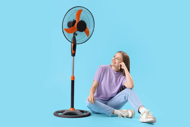 Young woman with electric fan sitting on blue background - Photo, Image