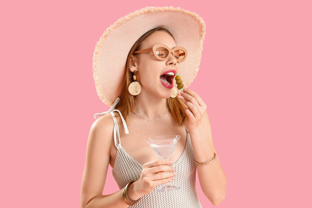 Belle jeune femme en osier chapeau avec verre de martini manger des olives sur fond rose - Photo, image
