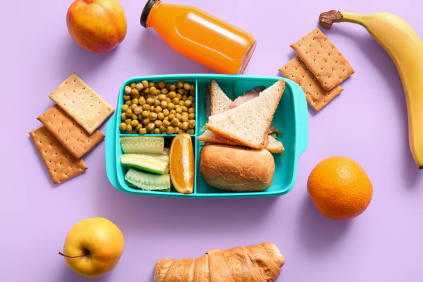 Lunchbox met heerlijk eten en flesje sap op lila achtergrond - Foto, afbeelding