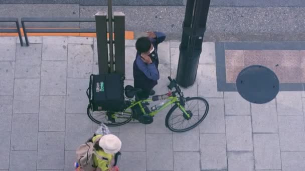 Tokyo, Japan-13 April, 2023: Slowmotion aerial view of a deliveryman waits on street standing next to Uber bike, ready to fulfill next order. A person in a delivery uniform with bicycle on sidewalk - Footage, Video