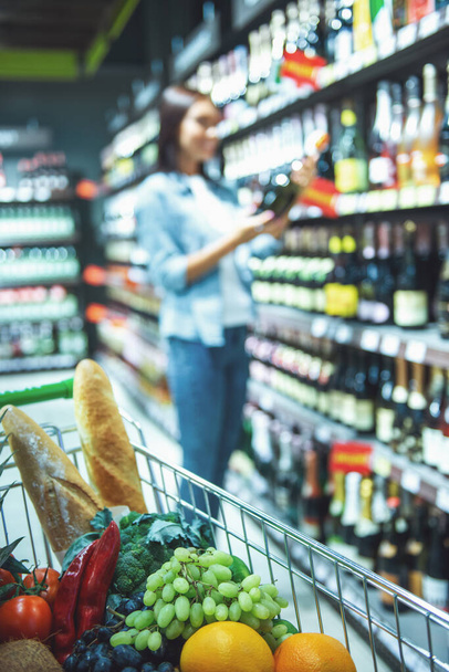A bevásárlókosár teljes élelmiszer, a háttérben a gyönyörű nő választotta egy ital a szupermarketben - Fotó, kép