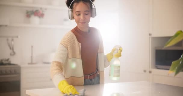 Een vrouw in een koptelefoon in de keuken schoonmaken met handschoenen, spray fles en glimlach, werken en luisteren naar streaming radio. Muziek, huishoudelijk werk en gelukkige huisvrouw met klusjes om stof of vuil af te vegen in huis - Video
