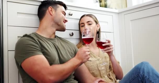 Couple on floor drinking red wine, cheers and smile to celebrate love, marriage and happy home. Toast with alcohol in glass, man and woman on ground to relax for conversation in kitchen together - Footage, Video