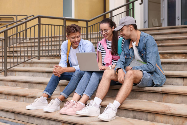 Happy young students studying together with laptop on steps outdoors - 写真・画像