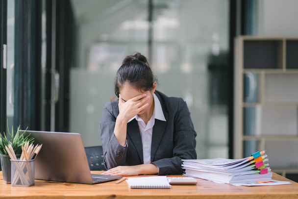 Die gestresste und erschöpfte asiatische Geschäftsfrau aus dem Millennium sitzt mit der Hand auf dem Kopf an ihrem Büroschreibtisch und deutet auf einen harten Arbeitstag hin, an dem sie mit Arbeit überlastet ist. - Foto, Bild